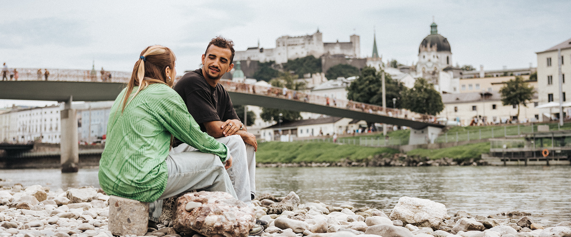 Eine Frau und ein Mann sitzen an der Salzach und unterhalten sich. 