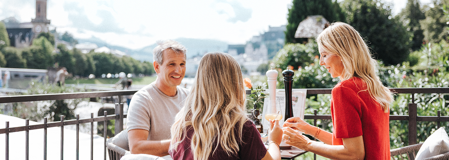 Eine Familie sitzt beim Frühstück in einem Kaffee an der Salzach mit Blick auf die Festung Hohensalzburg | © Niko Zuparic
