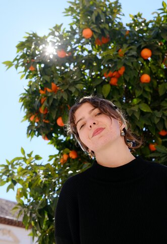 Die Musikerin Anna Gerstendorfer steht vor einem Orangenbaum und lächelt in die Kamera.  | © Anna Gerstendorfer